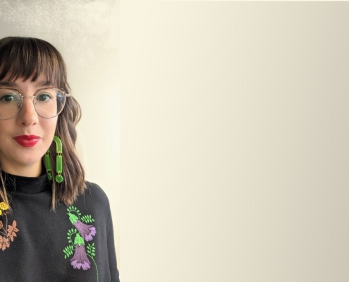 Hamon standing in front of a neutral background wearing a black mandarin top with flowers embroidered on it. Her brown hair is shoulder length. She is wearing green earrings that touch her shoulders and bright red lipstick.
