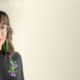 Hamon standing in front of a neutral background wearing a black mandarin top with flowers embroidered on it. Her brown hair is shoulder length. She is wearing green earrings that touch her shoulders and bright red lipstick.