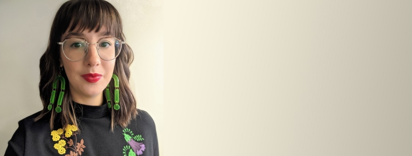 Hamon standing in front of a neutral background wearing a black mandarin top with flowers embroidered on it. Her brown hair is shoulder length. She is wearing green earrings that touch her shoulders and bright red lipstick.