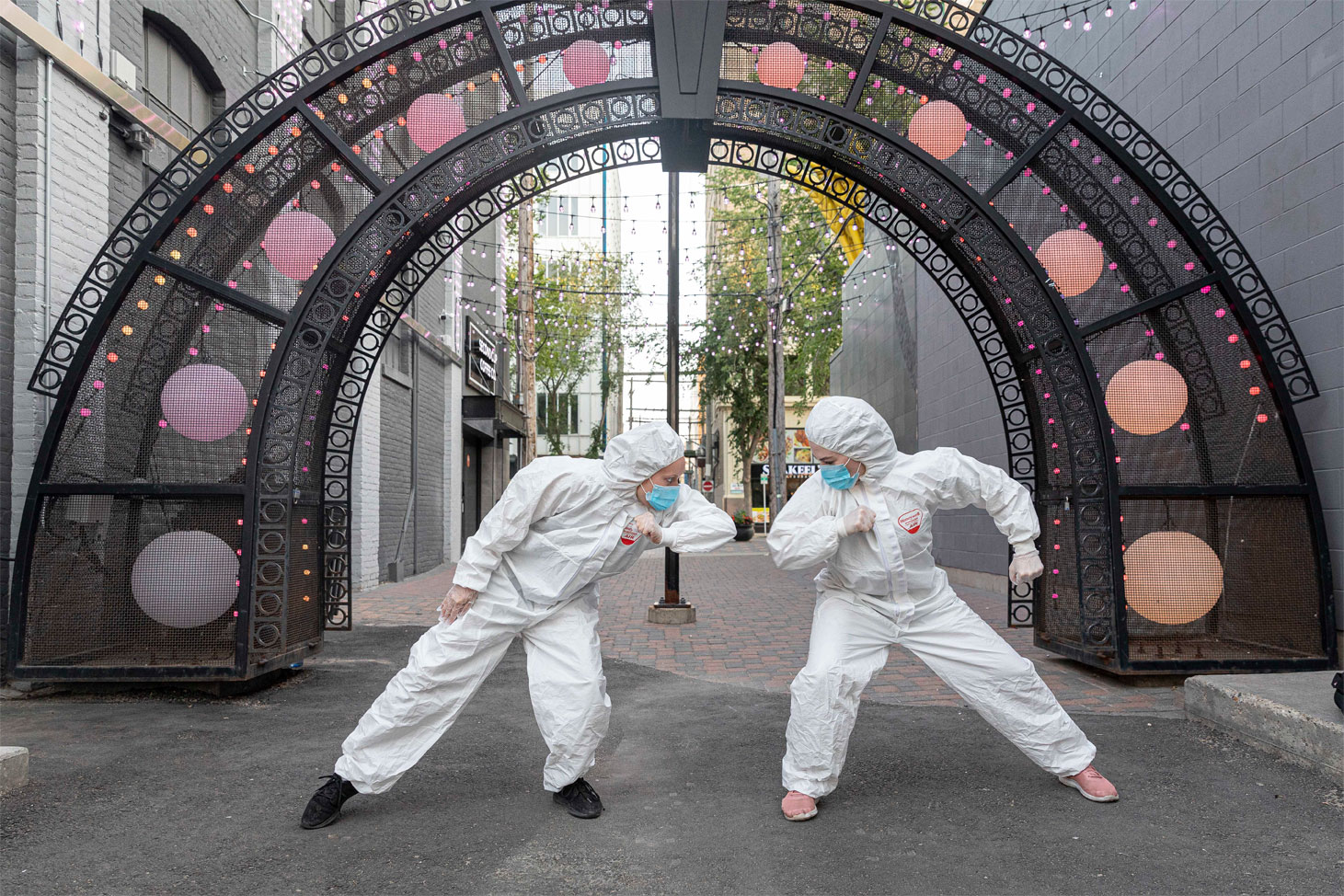 Two dancers in hazmat suits and masks face each other under an outdoor arch