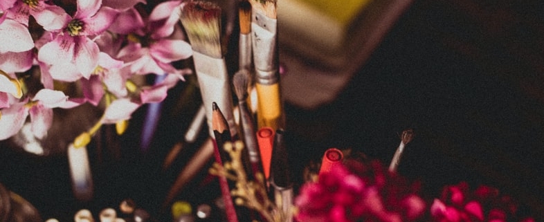 paint brushes in a cup beside a vase of flowers