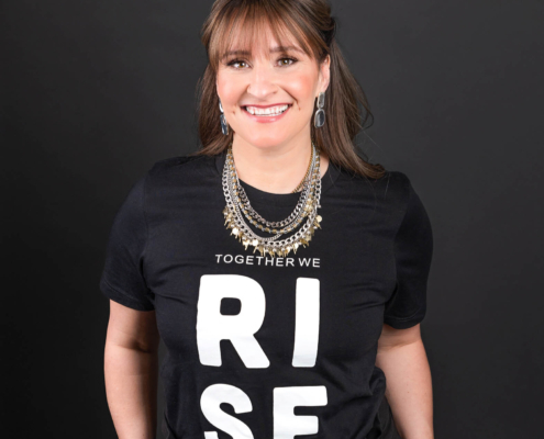 Jaimie in front of black background. Wearing a black shirt with white text reading Rise in all capital letters