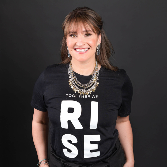 Jaimie in front of black background. Wearing a black shirt with white text reading Rise in all capital letters