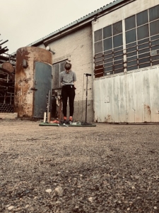 Edith standing in front of an industrial building - their head is down and they are standing by musical instruments and a microphone.