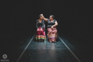 Two women sit side by side wearing ribbon skirts and black shirts in a black background.