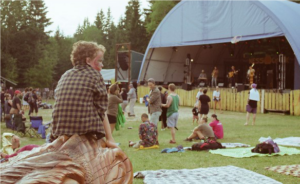 Boy sitting foreground while musicians on a stage perform in the background with scattered people in the background.