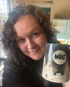 Krystal looking in to the camera - her curly brown hair shoulder length. She is holding a ceramic mug with WAC written on it.