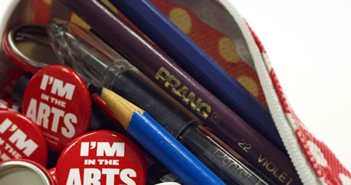 Open pencil case with pencil crayons falling out mixed with red buttons that read I'm in the Arts