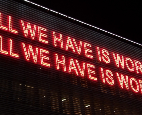 Red lighting against a black background reading "All we have is words. All we have is worlds"