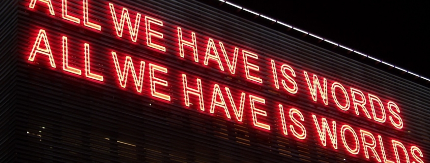 Red lighting against a black background reading "All we have is words. All we have is worlds"