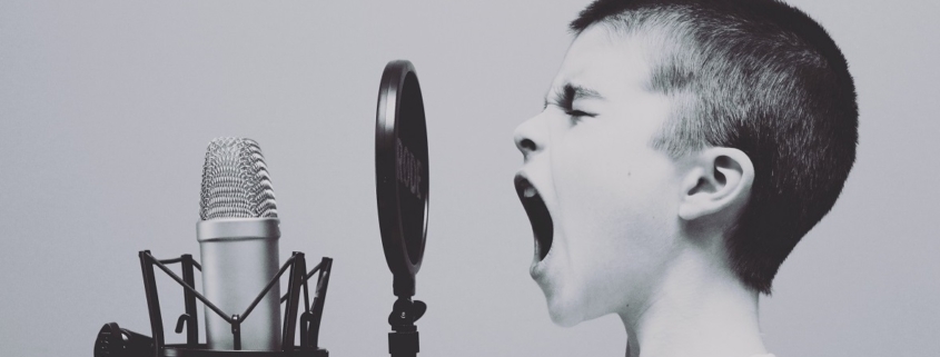 black and white image of boy with brush cut hair singing with mouth wide into a microphone.