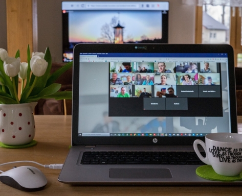 A laptop computer with a view of a Zoom meeting on the screen. There is a structure projected on a screen beyond the computer. There is a white and red polk a dot pitched with with tulips in it to the left of the computer and a white mug with a motivational phrase written on it in black sitting on the laptop.