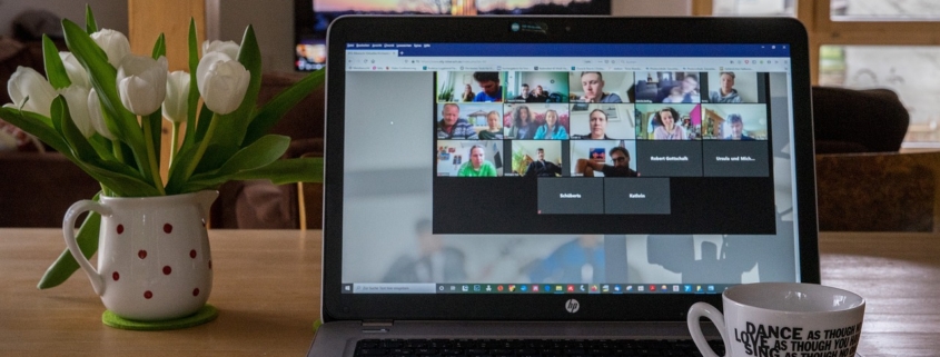 A laptop computer with a view of a Zoom meeting on the screen. There is a structure projected on a screen beyond the computer. There is a white and red polk a dot pitched with with tulips in it to the left of the computer and a white mug with a motivational phrase written on it in black sitting on the laptop.