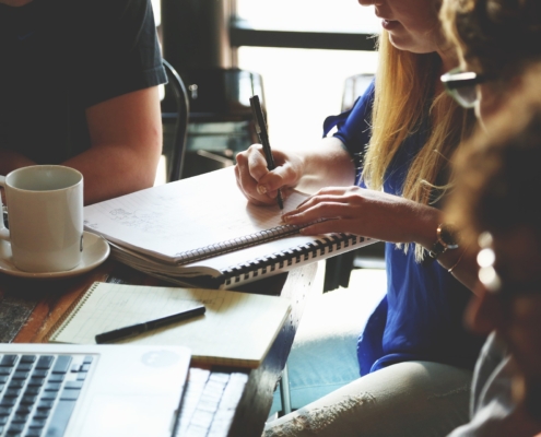 A group of four people sit around a table with a laptop, notebooks, pens and a coffee in a planning meeting