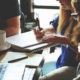 A group of four people sit around a table with a laptop, notebooks, pens and a coffee in a planning meeting