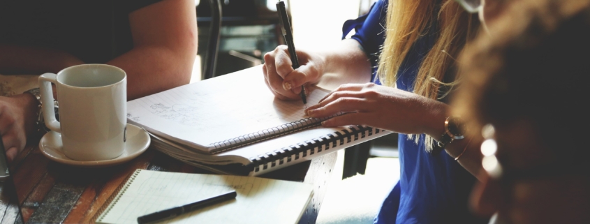 A group of four people sit around a table with a laptop, notebooks, pens and a coffee in a planning meeting