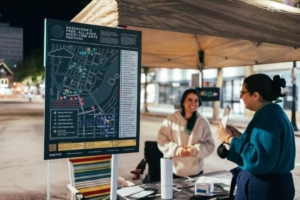 Two women in conversation by a map of Saskatoon featuring Nuit Blanche sites