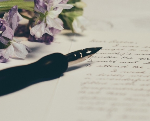 a fountain pen rests on a handwritten letter. There is purple and white flowers on the top left corner of the image.