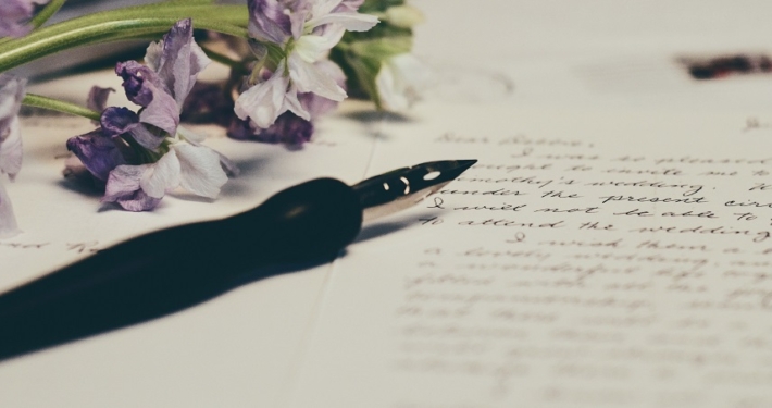 a fountain pen rests on a handwritten letter. There is purple and white flowers on the top left corner of the image.