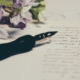 a fountain pen rests on a handwritten letter. There is purple and white flowers on the top left corner of the image.