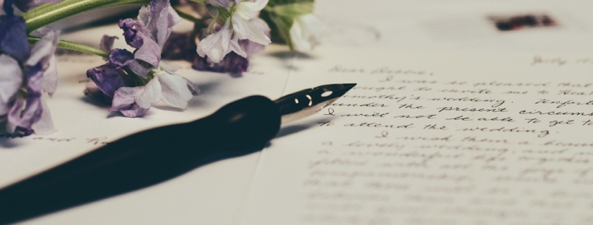 a fountain pen rests on a handwritten letter. There is purple and white flowers on the top left corner of the image.