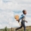 a boy walking across grass with a cloudy blue sky behind him. He is holding a ukelele.