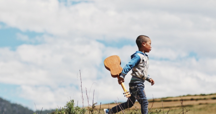 a boy walking across grass with a cloudy blue sky behind him. He is holding a ukelele.