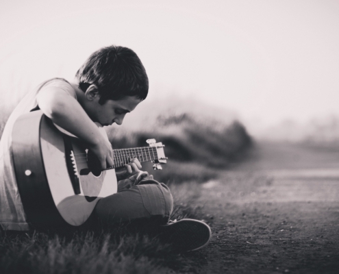 black and white photo of boy with short hair sitting cross legged on the ground playing a guitar.