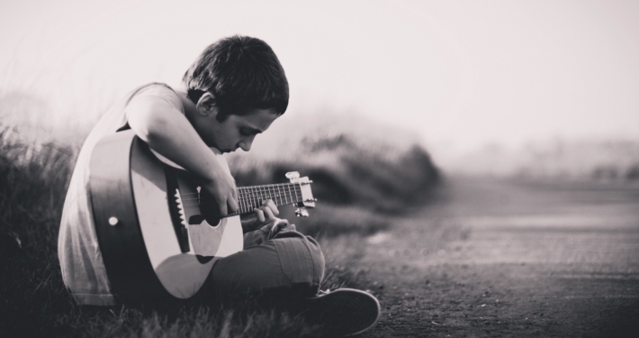 black and white photo of boy with short hair sitting cross legged on the ground playing a guitar.