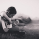 black and white photo of boy with short hair sitting cross legged on the ground playing a guitar.
