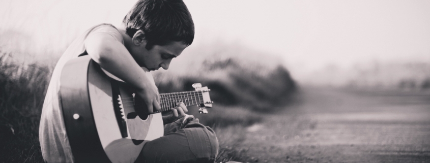 black and white photo of boy with short hair sitting cross legged on the ground playing a guitar.