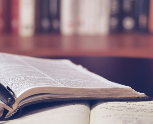 An open book with a pen in the spine on top of an open notebook. There is a full bookshelf in the background