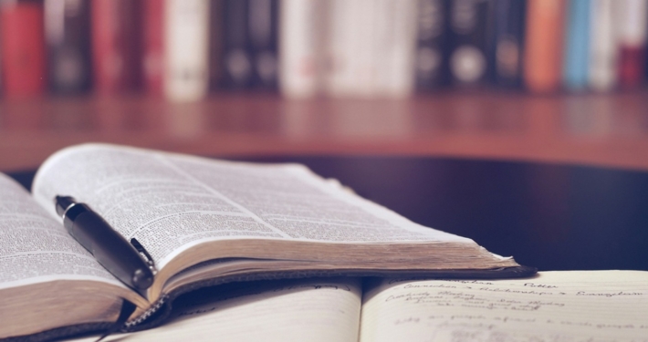 An open book with a pen in the spine on top of an open notebook. There is a full bookshelf in the background