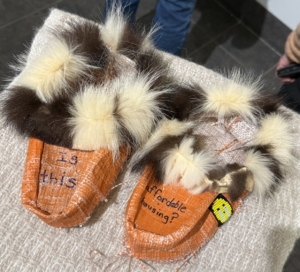 A pair of small handmade moccasins by Marcy Friesen constructed from orange tarps with fur trim and beadwork details. Text embroidered on the moccasins reads “Is this the best we can do?” and “Is this affordable housing?”, referencing housing precarity and care through traditional form.