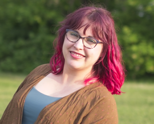 Woman with pink hair looking into camera, a smile on her face. She is wearing glasses