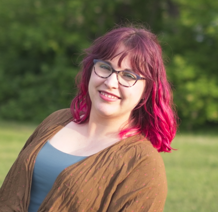 Woman with pink hair looking into camera, a smile on her face. She is wearing glasses