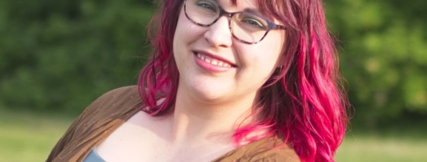 Woman with pink hair looking into camera, a smile on her face. She is wearing glasses