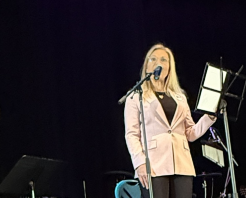two women stand against a black background. The woman in pink is speaking into a microphone while the other is interpreting what is said in sign language