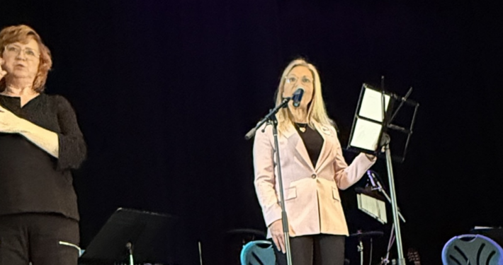 two women stand against a black background. The woman in pink is speaking into a microphone while the other is interpreting what is said in sign language