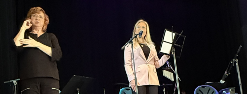 two women stand against a black background. The woman in pink is speaking into a microphone while the other is interpreting what is said in sign language
