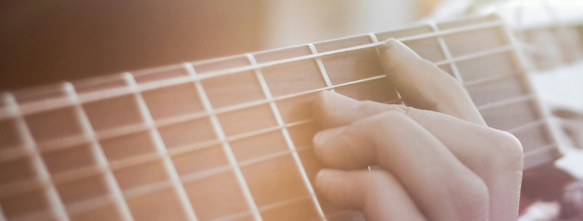 Close-up of the neck of a guitar with a hand placed on a chord. Sun shining down on both