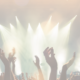 A crowd of concert goers with their arms in the air. In front of a brightly lit stage - too bright to see what is on the stage